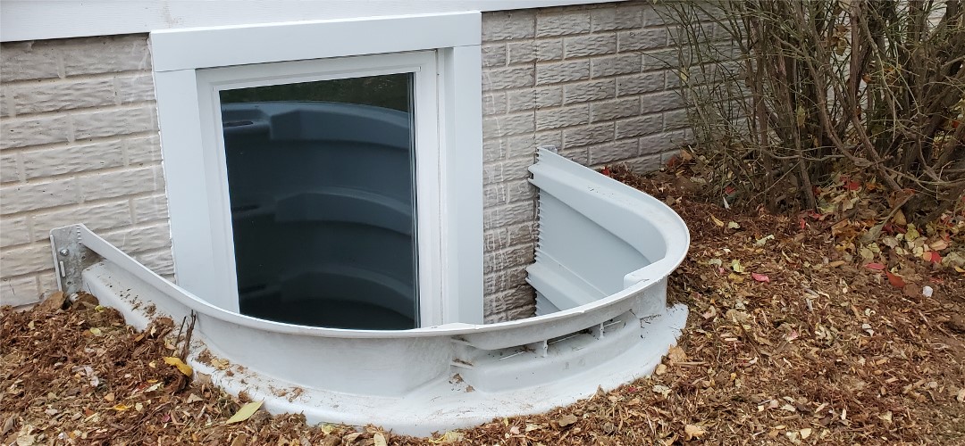 A white window well with a leaf covered ground.